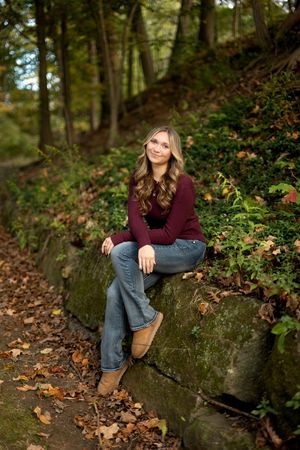Smiling young woman in burgundy sweater and jeans sitting on a stone wall in autumn forest.