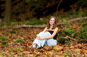 Smiling teenage girl sitting in autumn leaves on a forest floor surrounded by fall foliage.