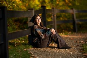 Teenage girl with glasses sitting against a wooden fence on a gravel path surrounded by fall foliage.