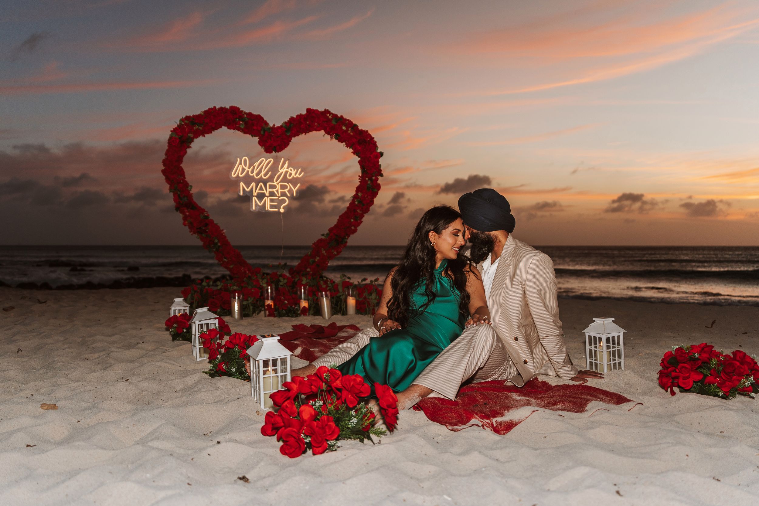 Couple holding hands at sunset beach proposal setup with floral arch, candles, and Marry Me sign on sandy shore.
