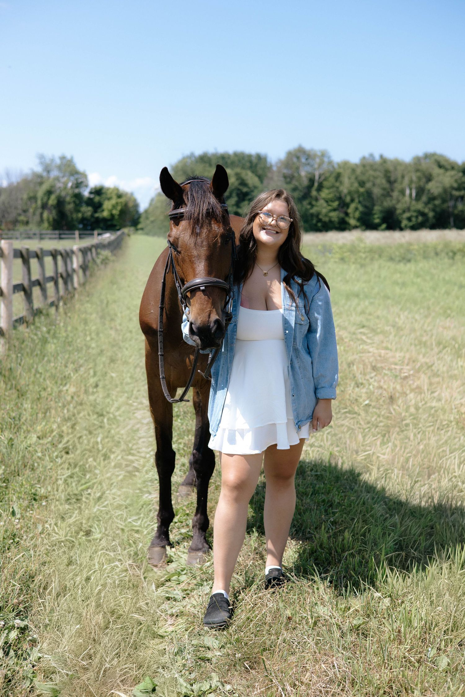 Smiling girl in white dress and denim jacket walking a bay horse along a sunny fenced field.