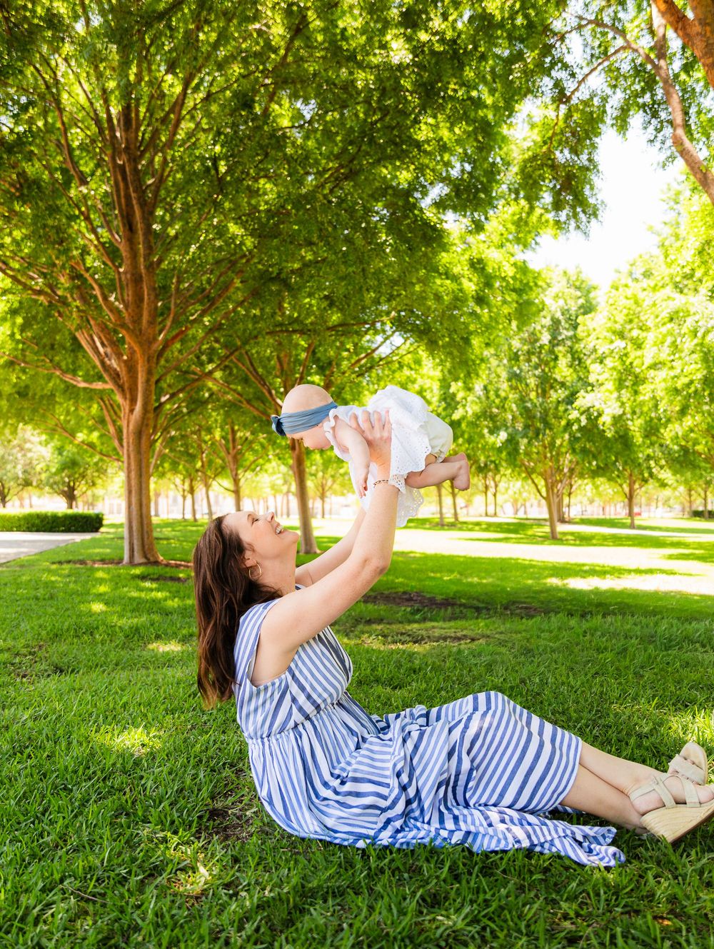 A mother in a striped dress lies on green grass in a park, joyfully lifting her baby up in the air.