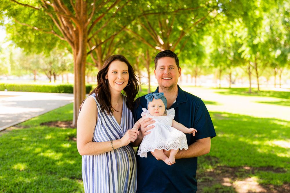 A smiling couple stands outdoors in a sunny park holding their baby dressed in white, surrounded by lush green trees.