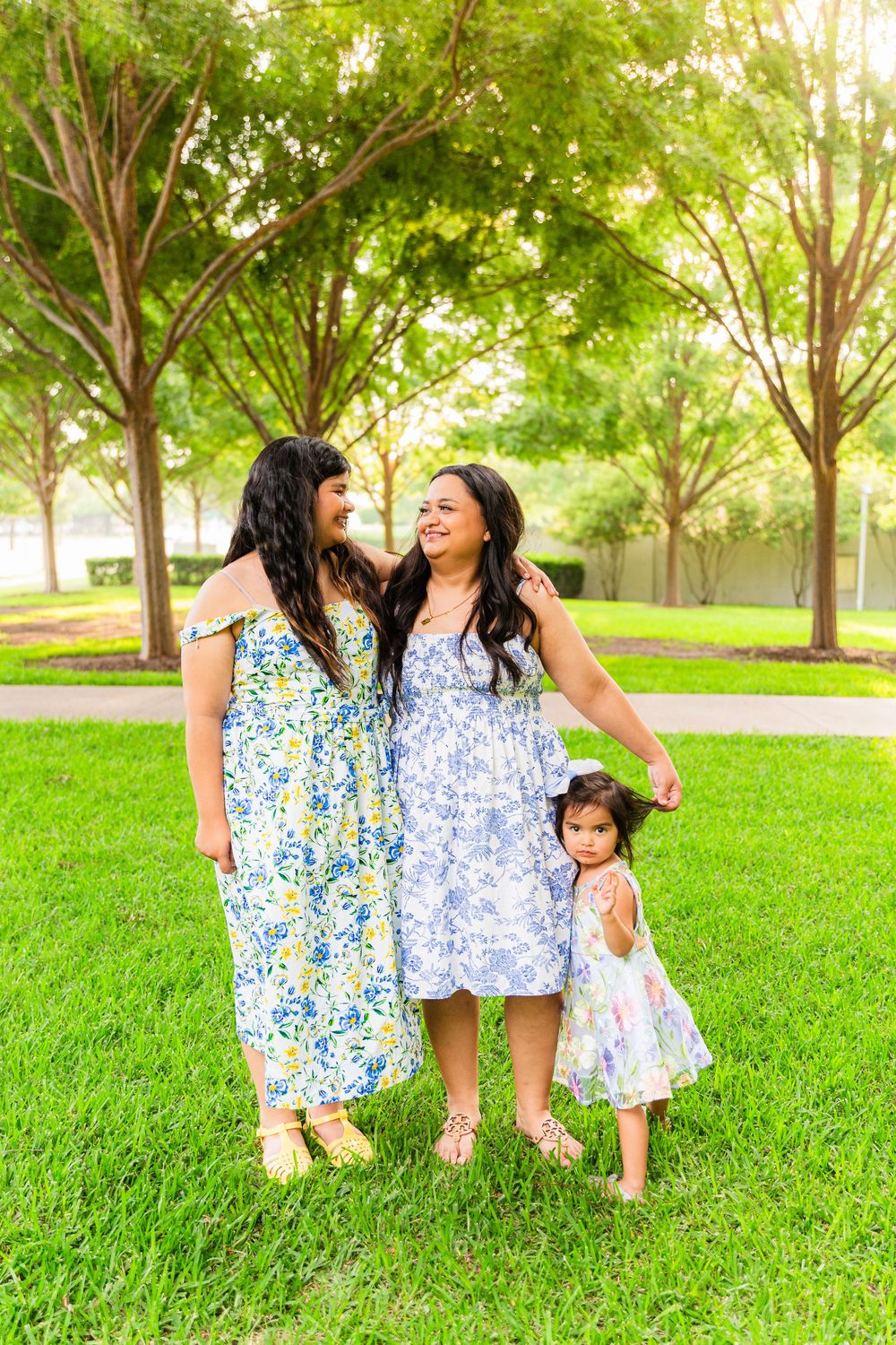Two women and a toddler girl in floral dresses pose together on green grass in a sunny park.