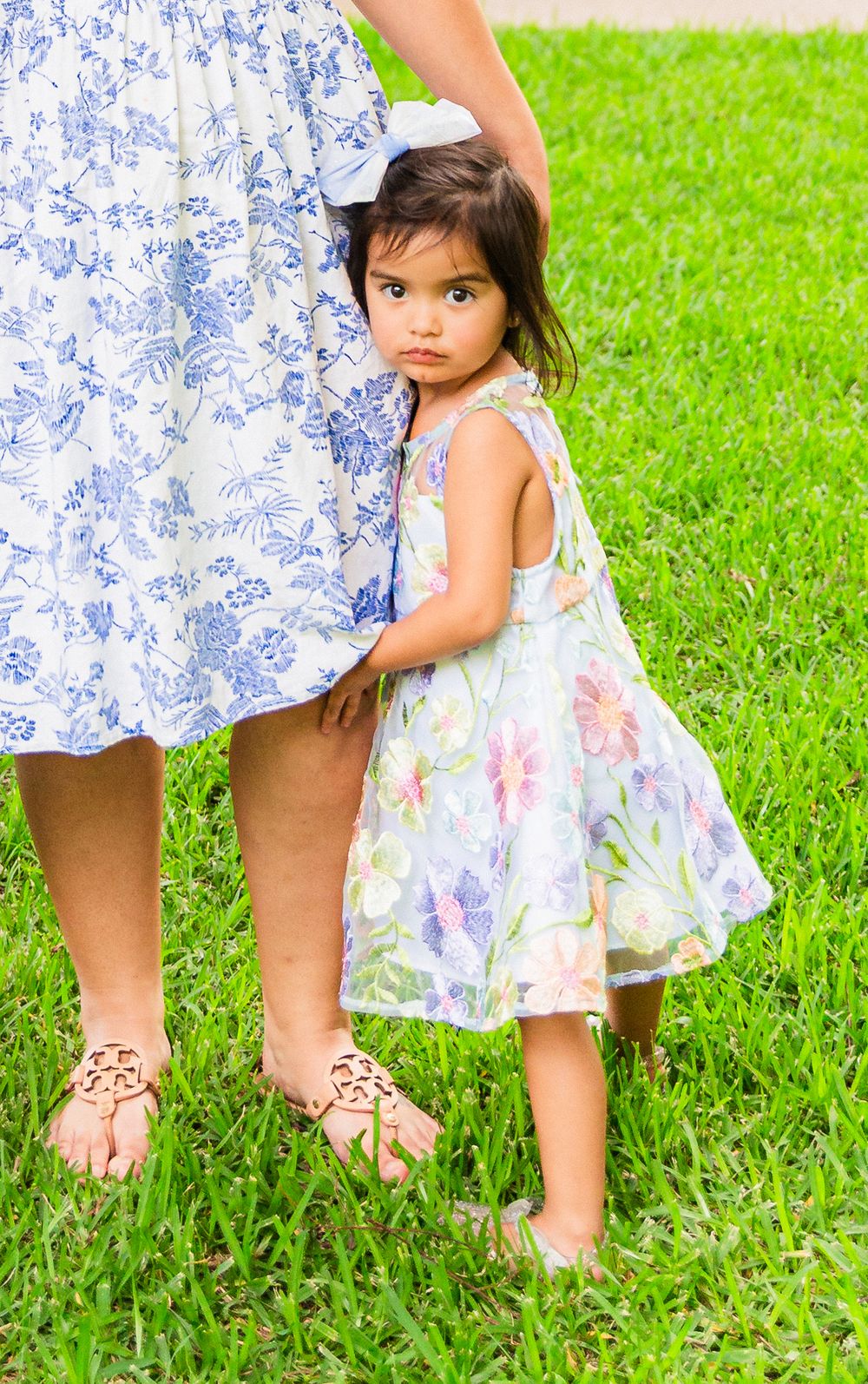A young girl in a floral dress clings to her mother's leg while standing on green grass outdoors.