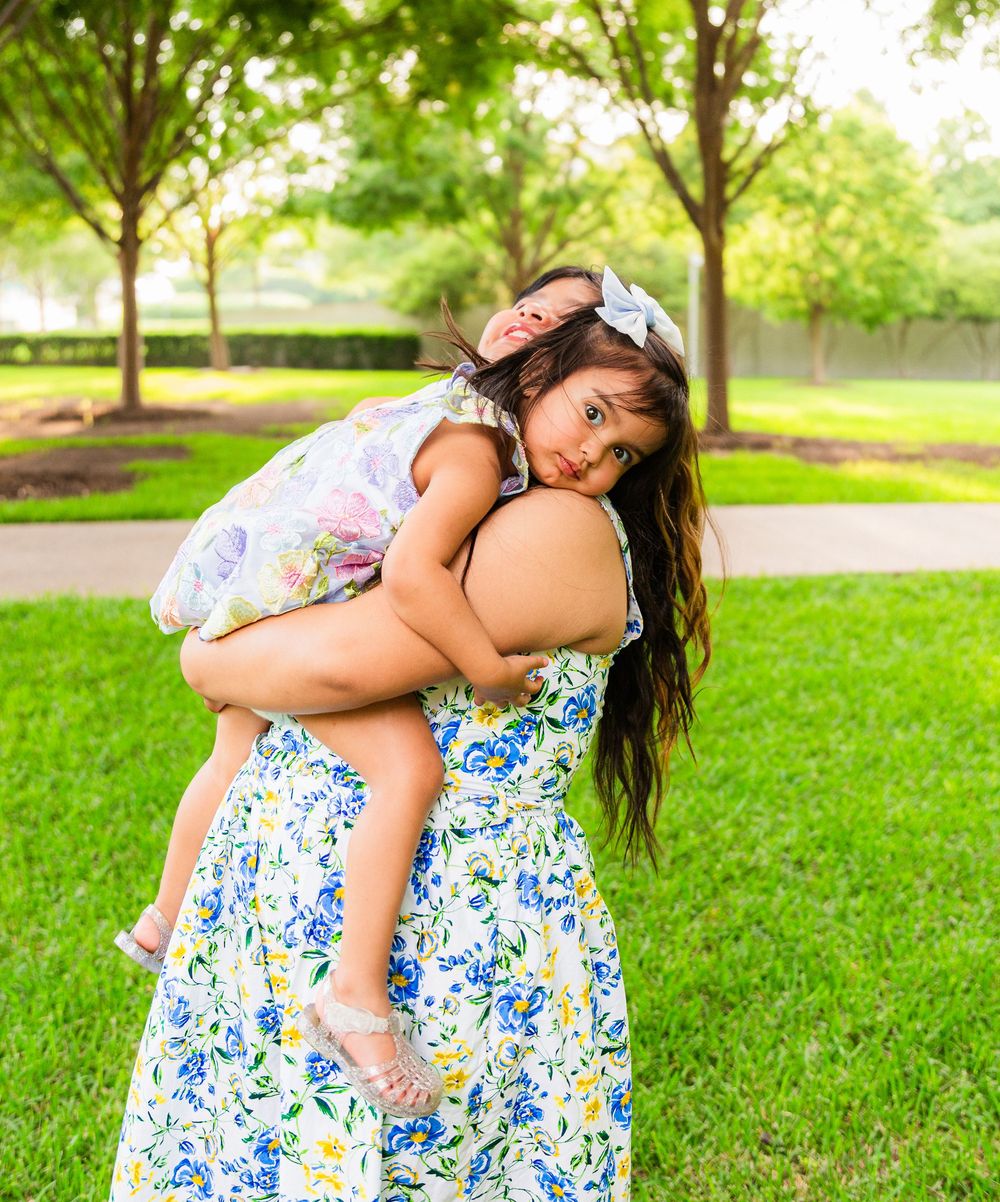 A woman in a floral dress holds a young girl in a park with lush green grass and trees.