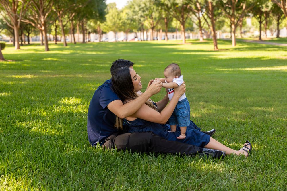 A mother sits on green grass in a sunny park, playfully holding her baby upright while smiling lovingly.