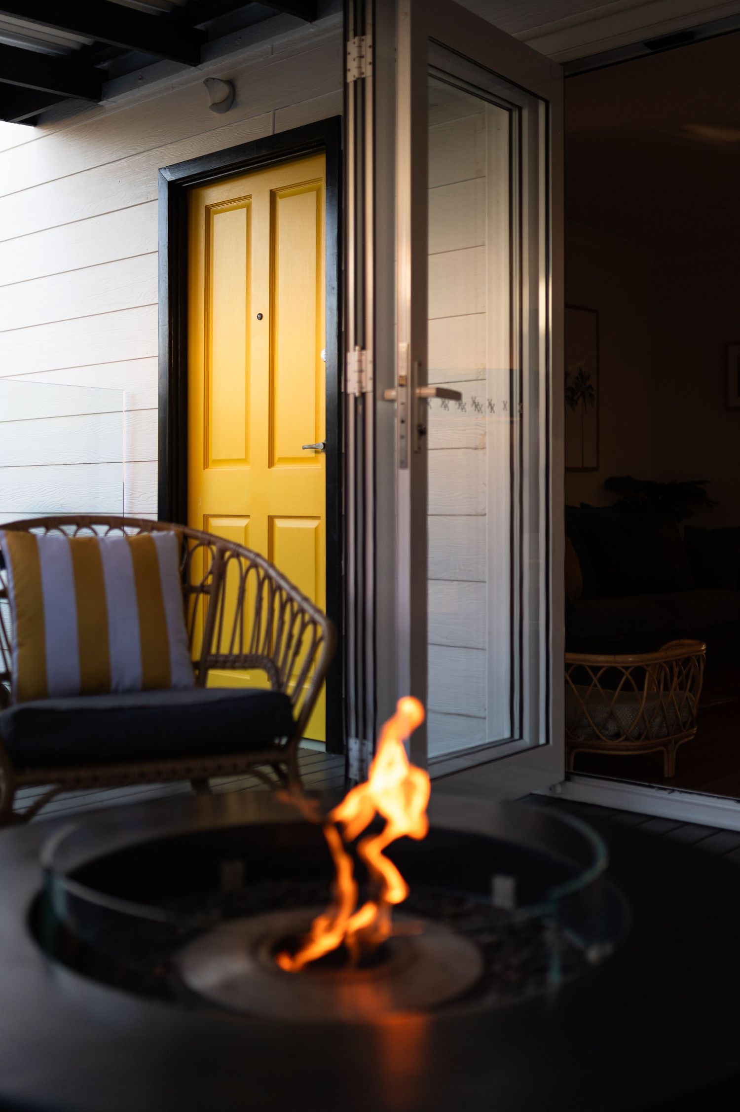 Cozy outdoor patio with a glowing fire pit, rattan chairs, and a vibrant yellow door on a white building at dusk.