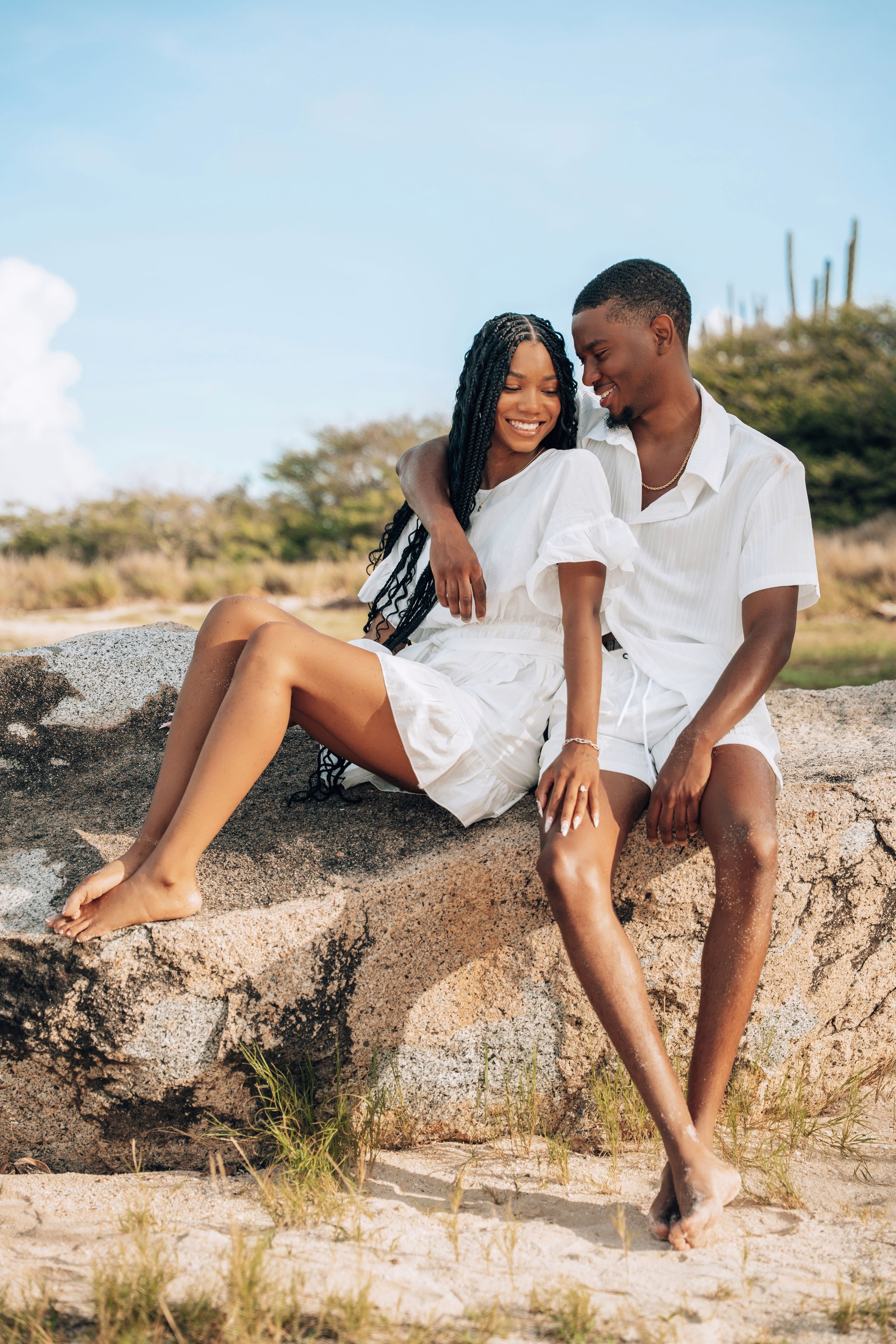 A happy Black couple in matching white outfits sitting on rocks outdoors, smiling and enjoying a sunny day together.