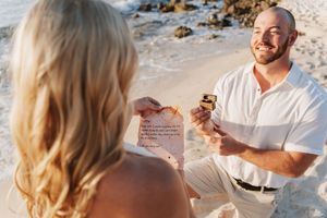 Man kneeling on beach proposing with ring box and love letter to blonde woman at sunset.