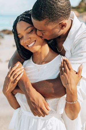 A happy couple embraces on a beach, the man kissing the woman's cheek as she smiles, both wearing white outfits.