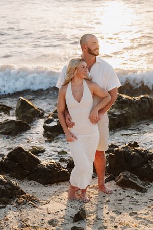Couple embracing on rocky beach at golden sunset, woman in white dress, man in casual white shirt and shorts.