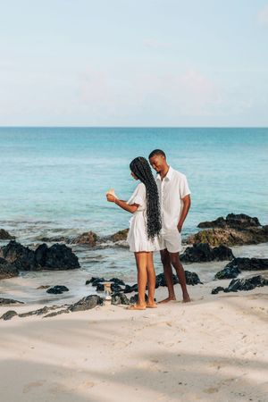A couple in white outfits stands on a tropical beach with turquoise water and rocky shores.