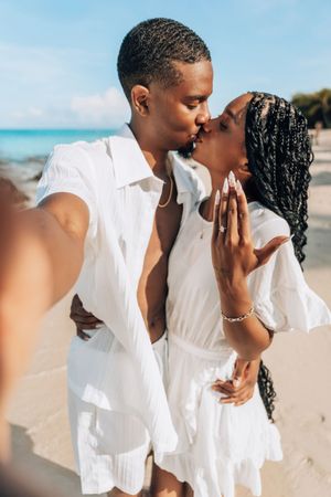 A couple in white outfits sharing a kiss on a sunny beach while taking a selfie.