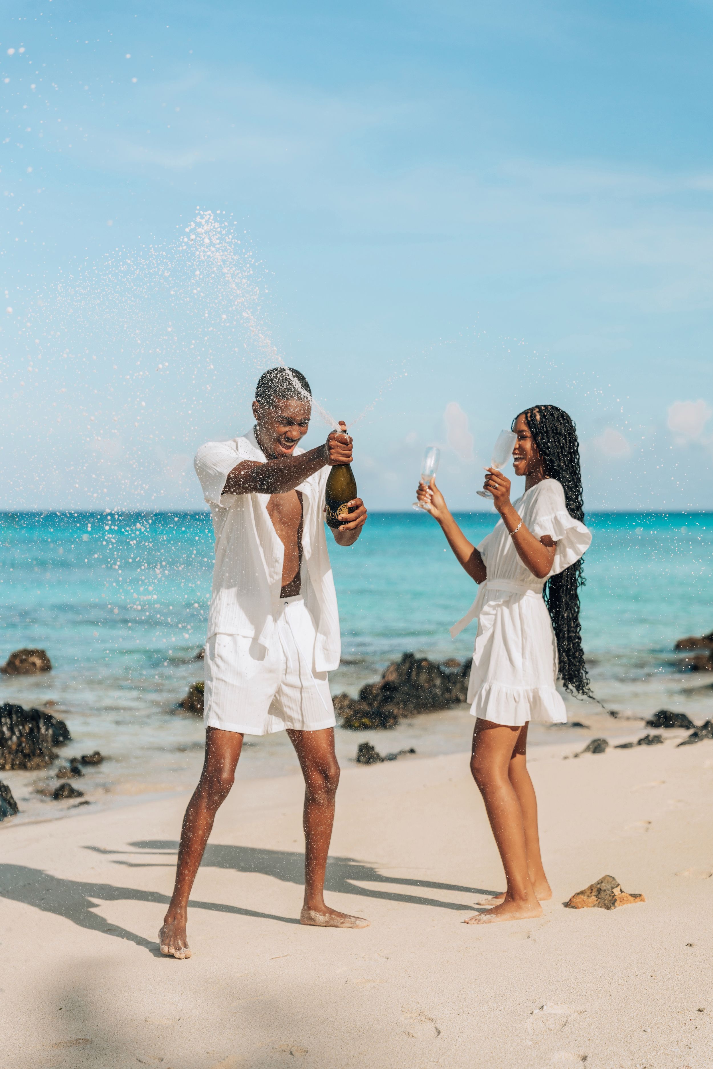 Couple in white outfits celebrating with champagne on a tropical beach with turquoise water.