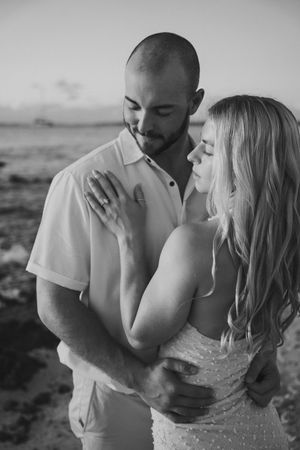 Black and white photo of a couple embracing on a beach, woman in white dress, man in white shirt, romantic sunset backdrop.