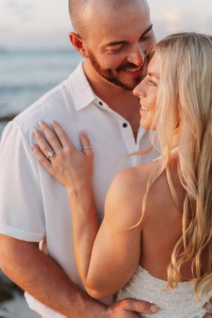 A smiling couple embraces on a beach at sunset, woman showing engagement ring in a romantic moment.