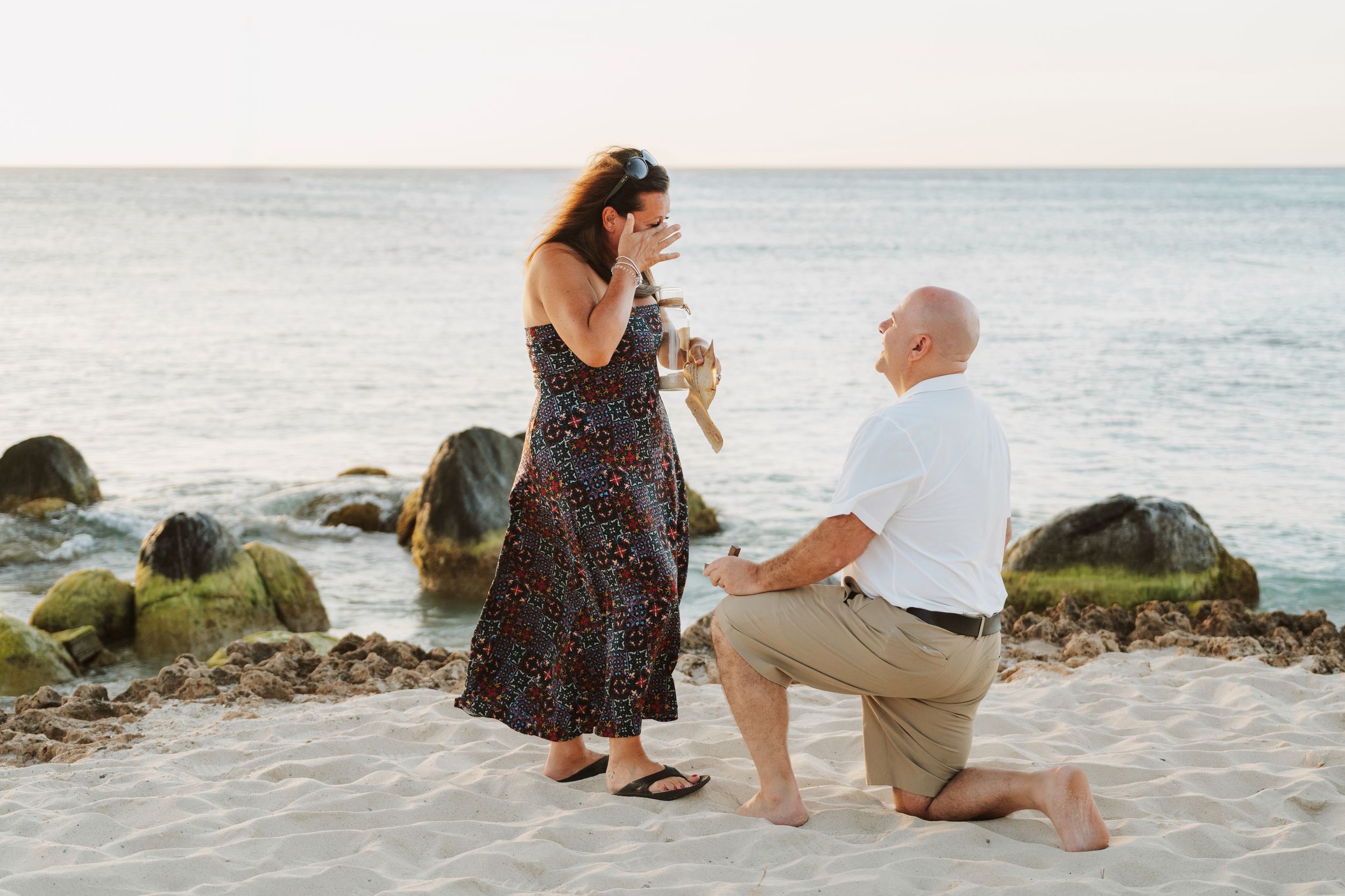 Man proposing on one knee to woman on sandy beach at sunset, with rocky shoreline and calm ocean in background.