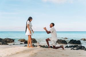 Man proposing to woman on a tropical beach, kneeling with ring while waves and rocks create a romantic backdrop.