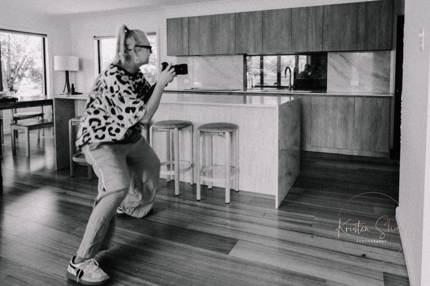 Black and white photo of a photographer crouching to take a picture inside a modern kitchen with wooden floors.