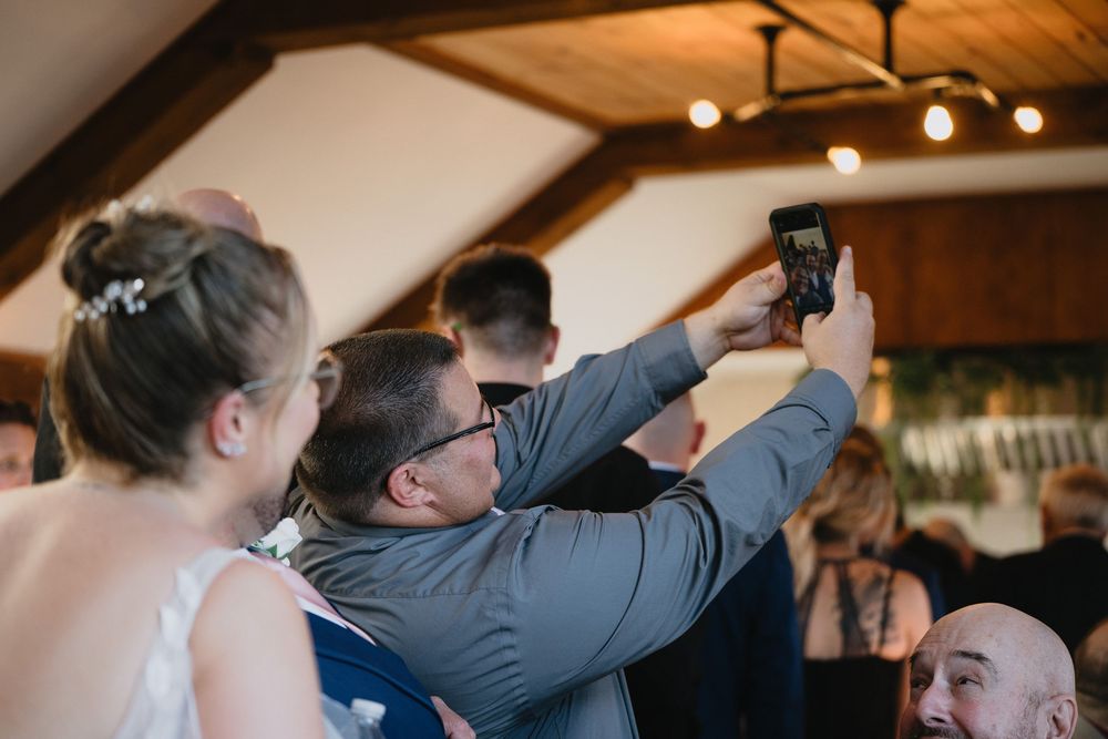 Wedding guests taking a selfie with a smartphone at an indoor reception venue with wooden beam ceiling.