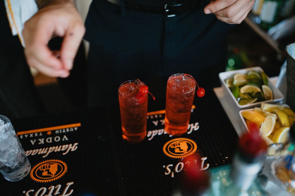 Two red cocktails on a Tito's Vodka bar mat, garnished with cherries, with citrus fruits nearby.