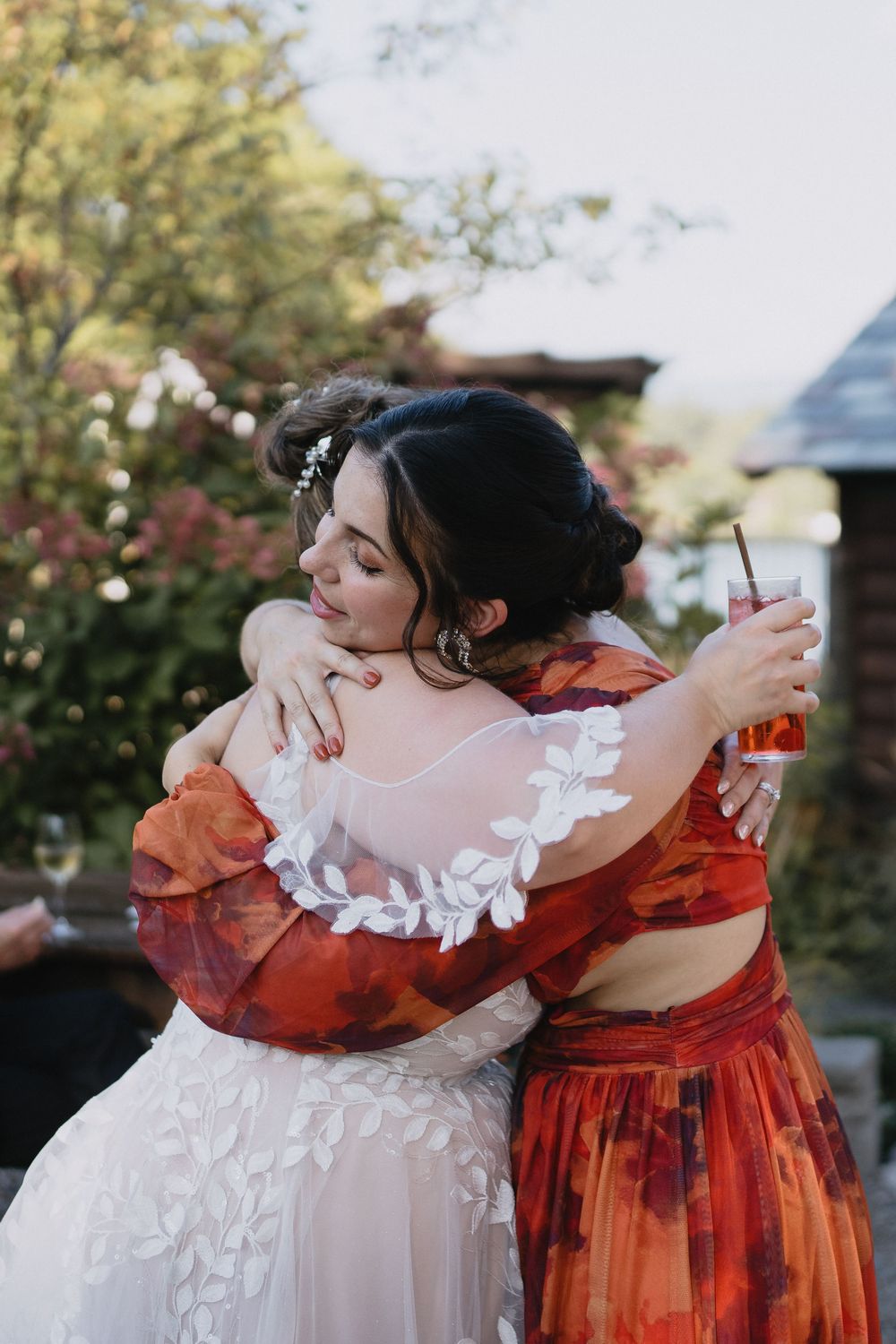 Two women share a warm embrace at a wedding, one in a white gown and the other in a red floral dress.
