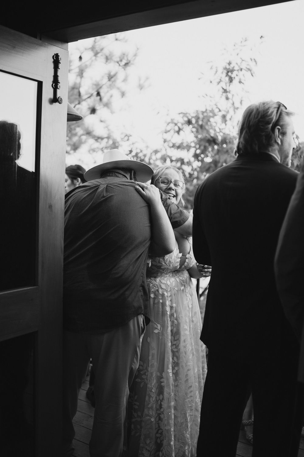Black and white wedding photo of guests embracing near a doorway, woman in lace gown and man in cowboy hat.