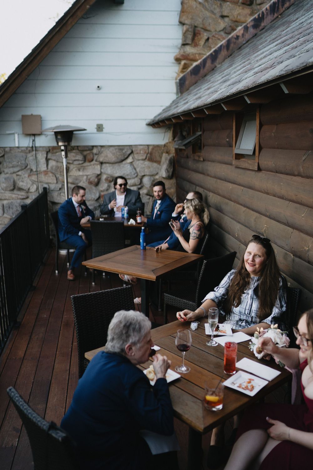Groups of people dining outdoors on a rustic wooden deck patio beside a log cabin building.