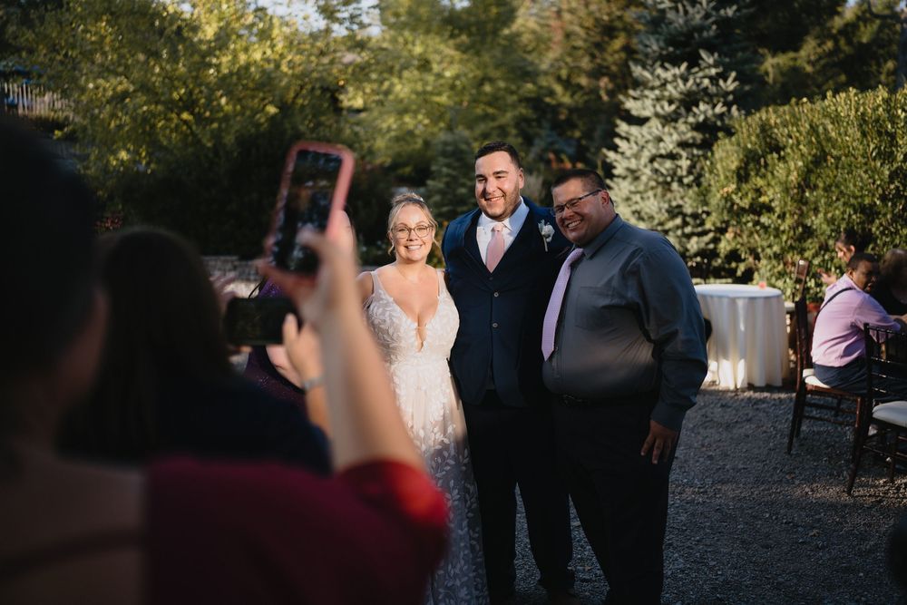 A bride and groom pose for photos with a guest outdoors at a wedding reception surrounded by lush greenery.