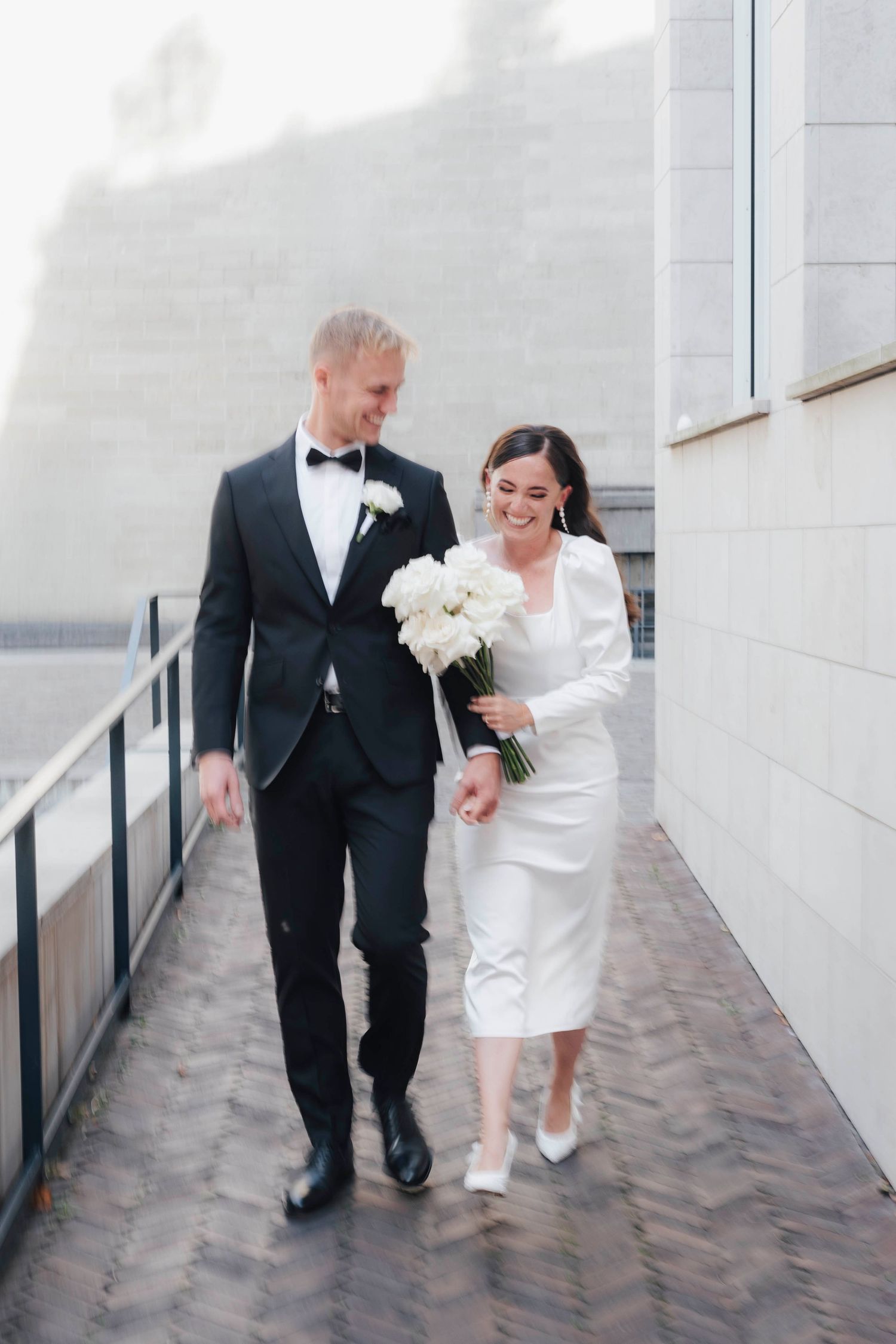Bride and groom walking together outdoors, she holds white bouquet in midi dress, he wears black tuxedo.