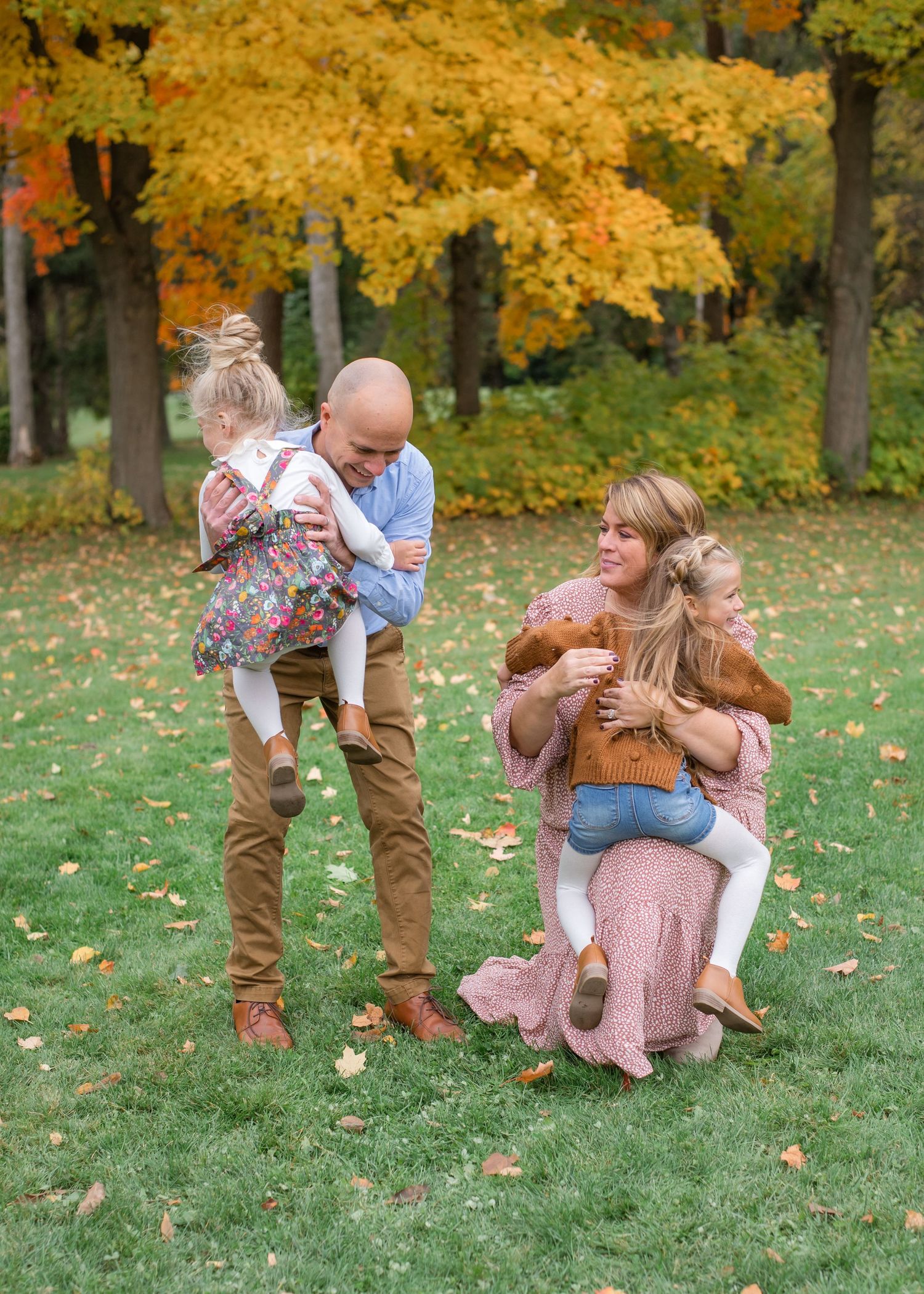 Family of four enjoying autumn outdoors, parents holding young children among vibrant fall foliage on green grass.