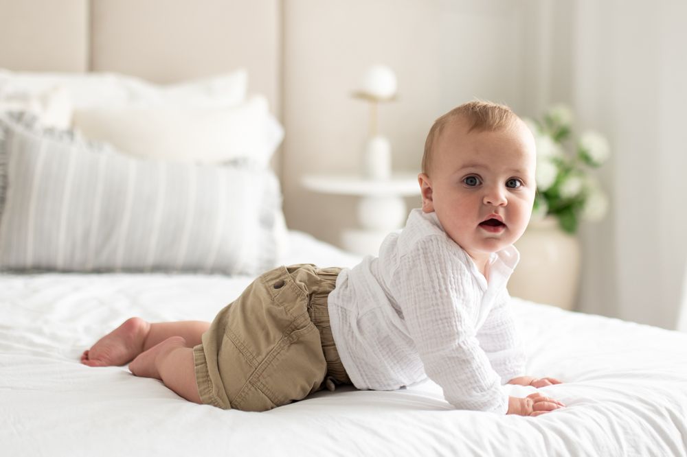 Adorable baby in white top and khaki shorts crawling on a white bed in a bright, modern nursery.