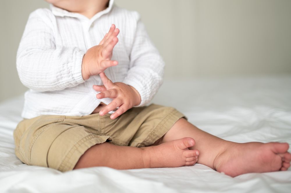 Adorable baby in white top and khaki shorts sitting on bed, clapping hands.