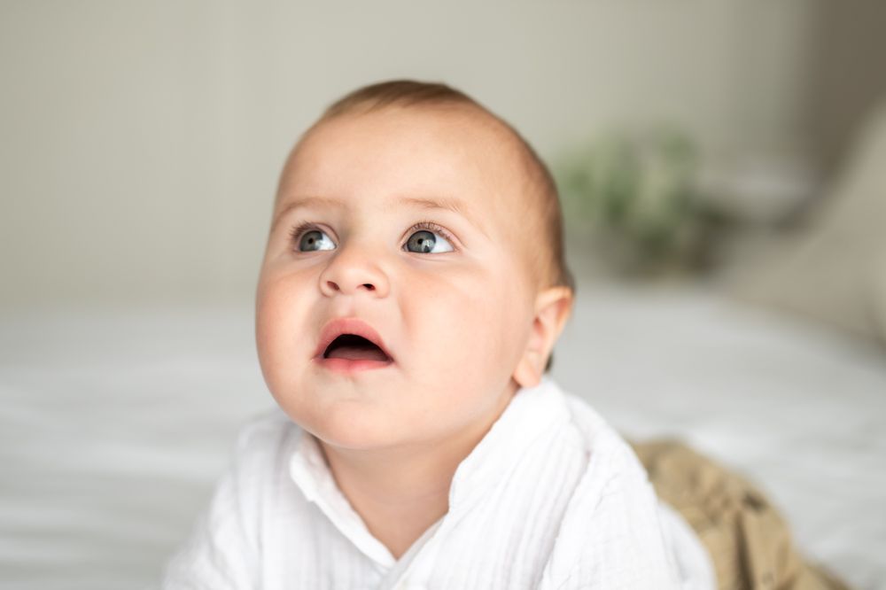 Adorable baby in white outfit lying on tummy, looking up with curious blue eyes in bright minimalist room.