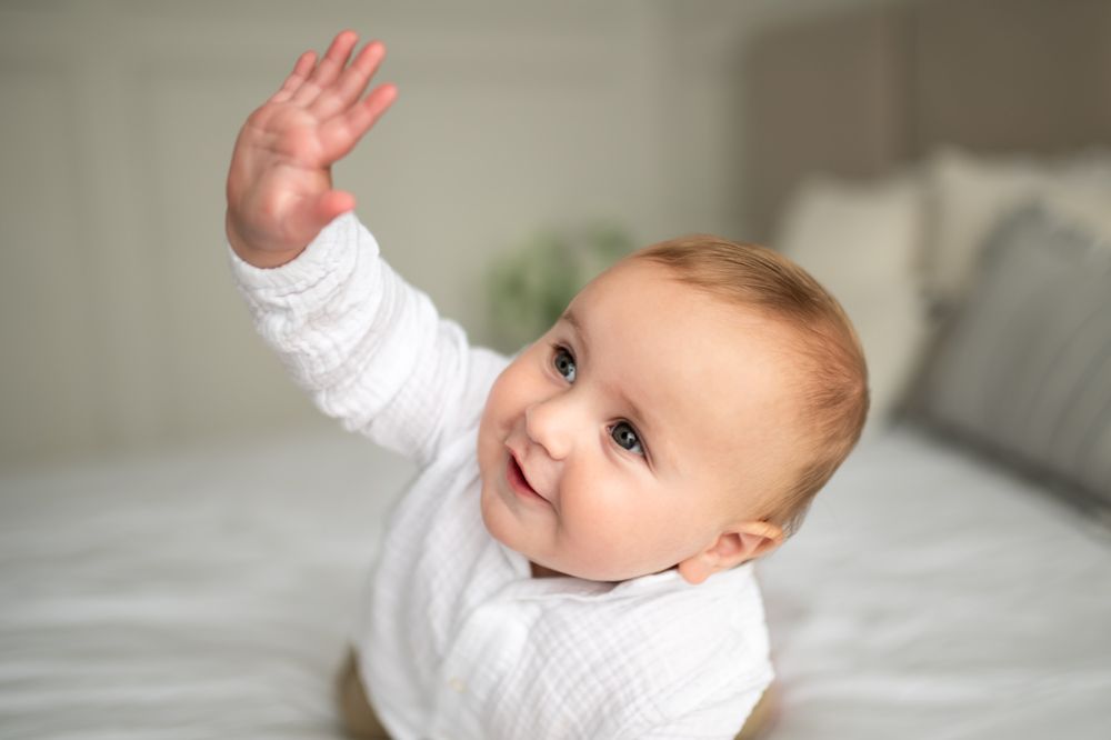 Adorable baby in white outfit raising hand while sitting on a bed.