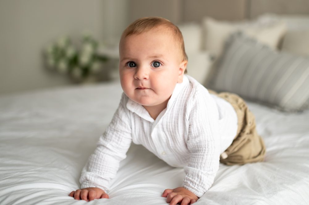 Adorable baby in white knit outfit doing tummy time on a white bed.