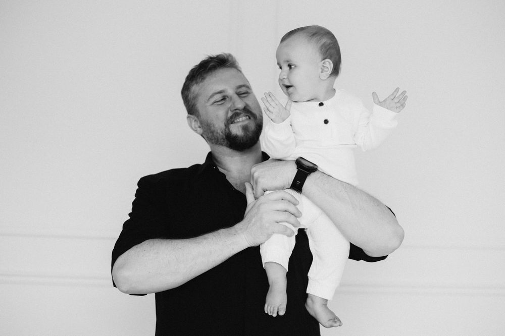 Black and white photo of a bearded man smiling while holding a happy baby dressed in white.