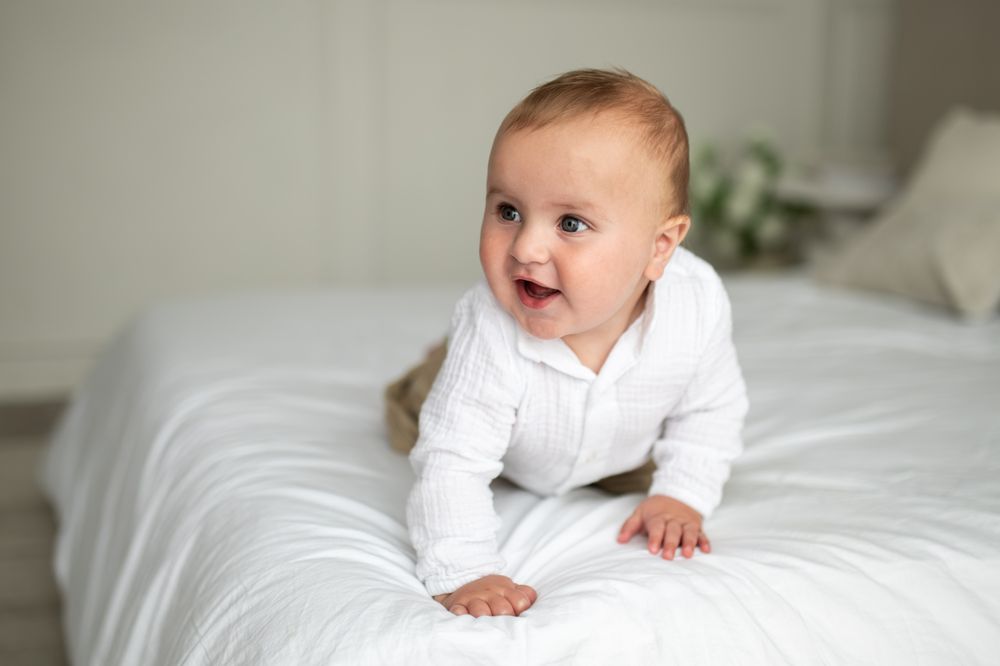 Smiling baby in white onesie doing tummy time on a white bed.