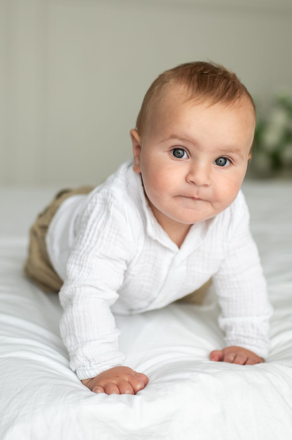 Cute baby in white shirt on bed, looking at camera while doing tummy time.
