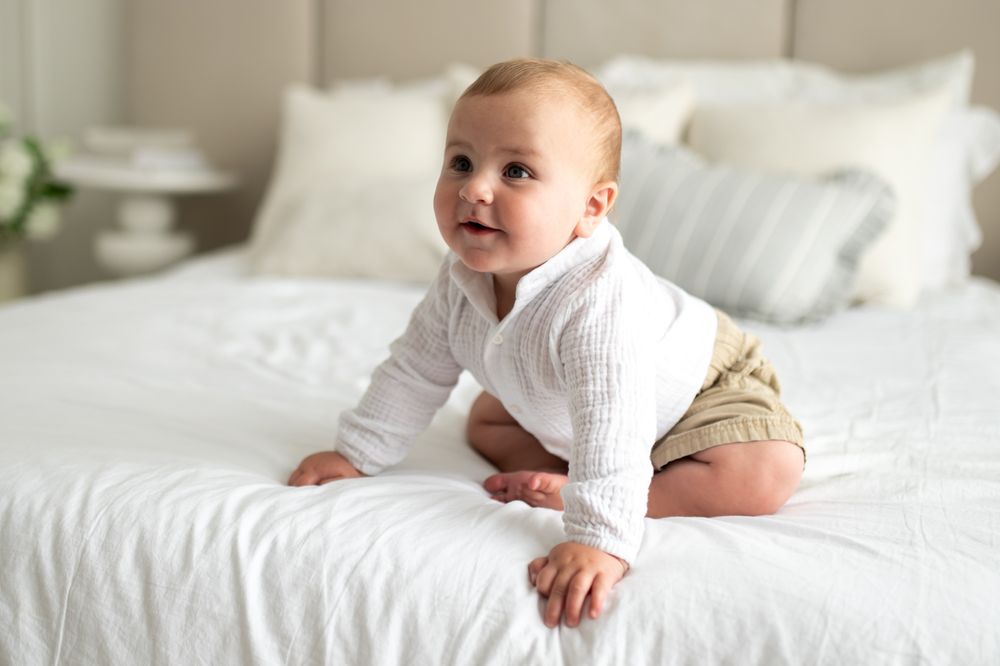 Adorable baby in white shirt and khaki shorts crawling on a white bed, looking up with a curious smile.
