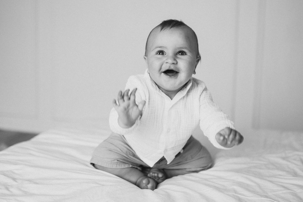 Black and white photo of a happy smiling baby sitting up on a bed, waving hand at camera.
