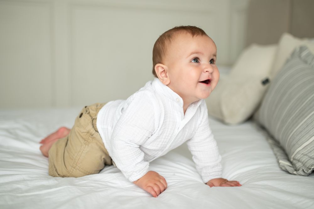 Happy baby in white top and tan pants crawling on a white bed.