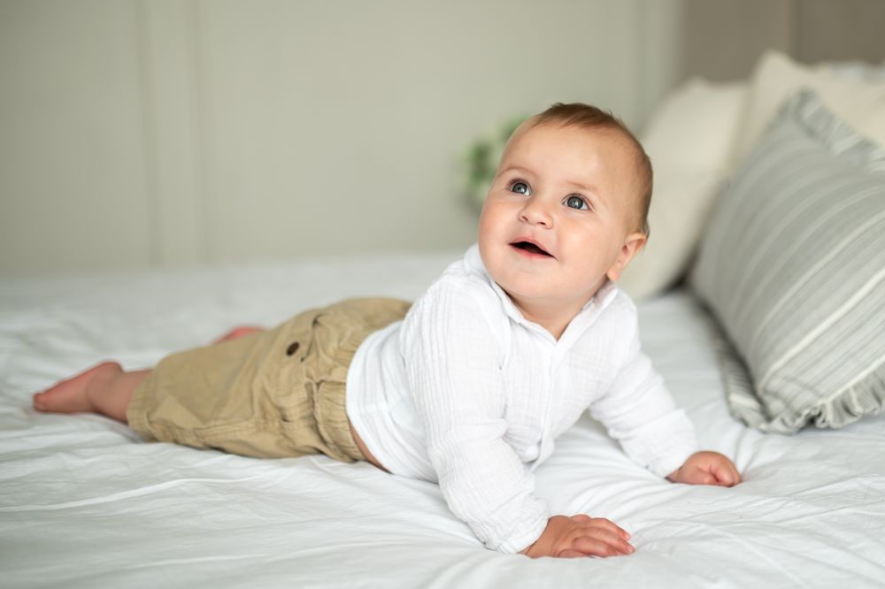 Smiling baby in white onesie and khaki pants doing tummy time on a white bed with gray pillows.