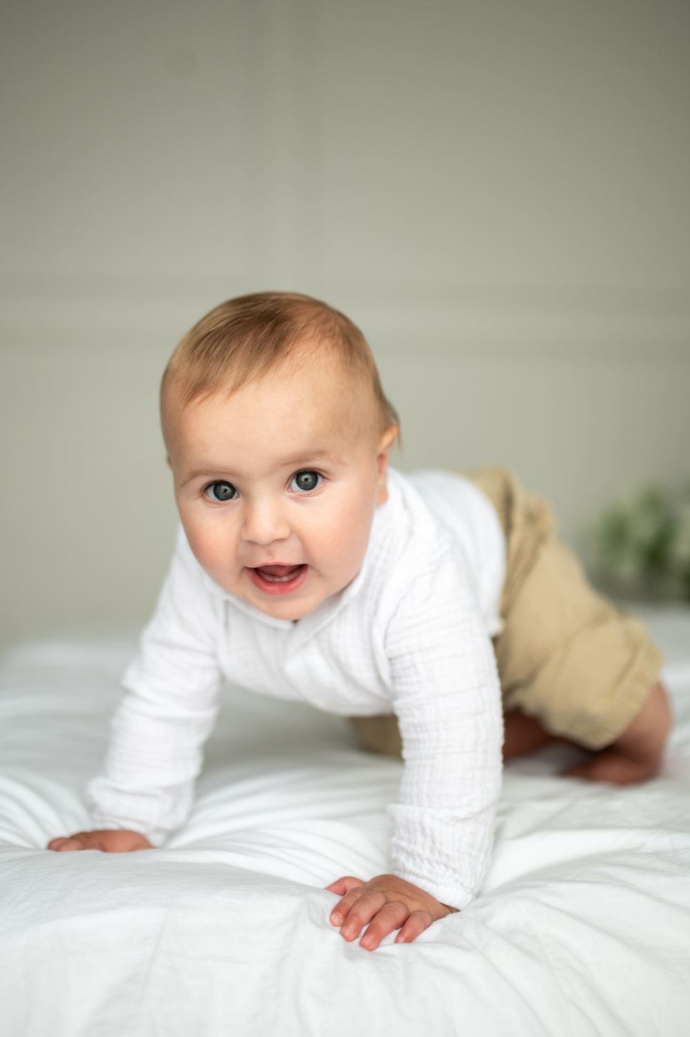 Happy baby in white onesie and tan pants crawling on white bedding, smiling at the camera.