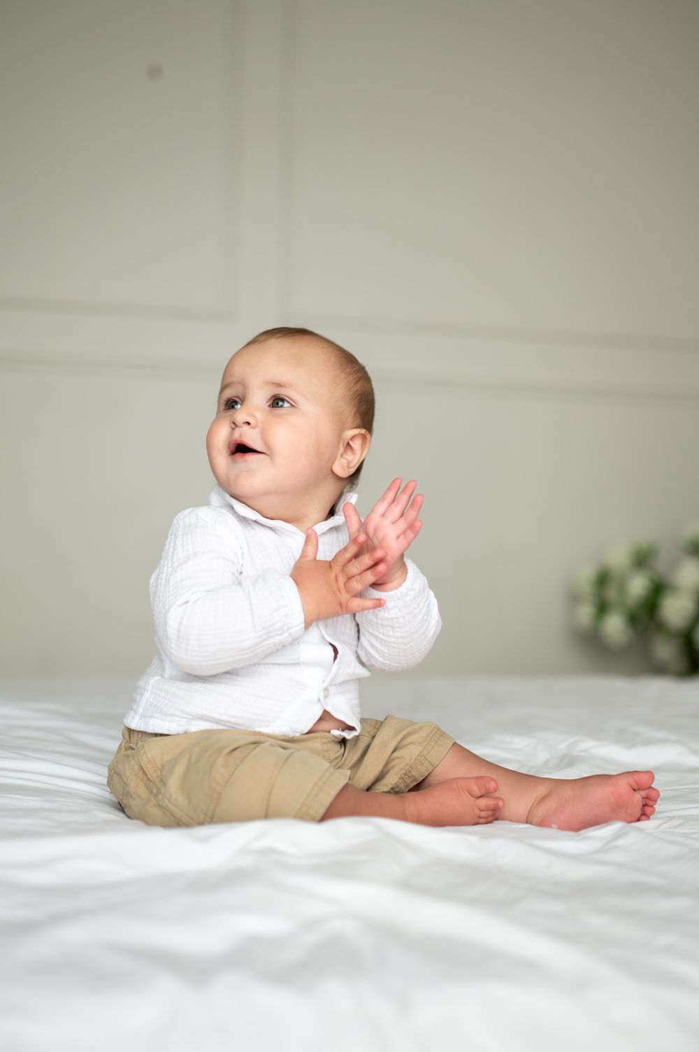 A happy baby in a white top and khaki pants sits on a white bed, clapping hands and looking up.