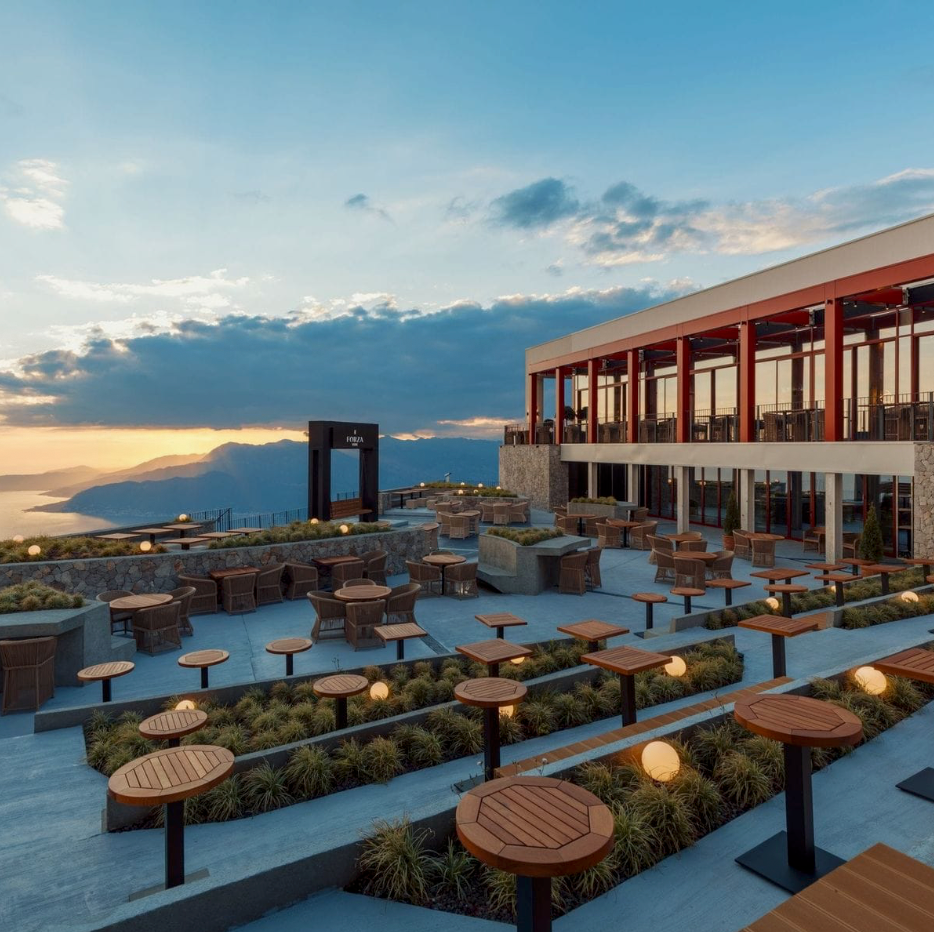 Rooftop terrace with wooden tables and chairs overlooking a scenic coastal sunset view at a modern resort.