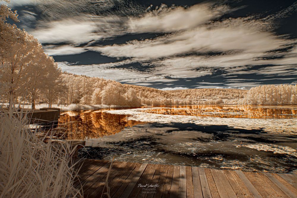 Infrared landscape photo of a serene lake reflecting golden trees under dramatic cloudy skies with a wooden deck foreground.
