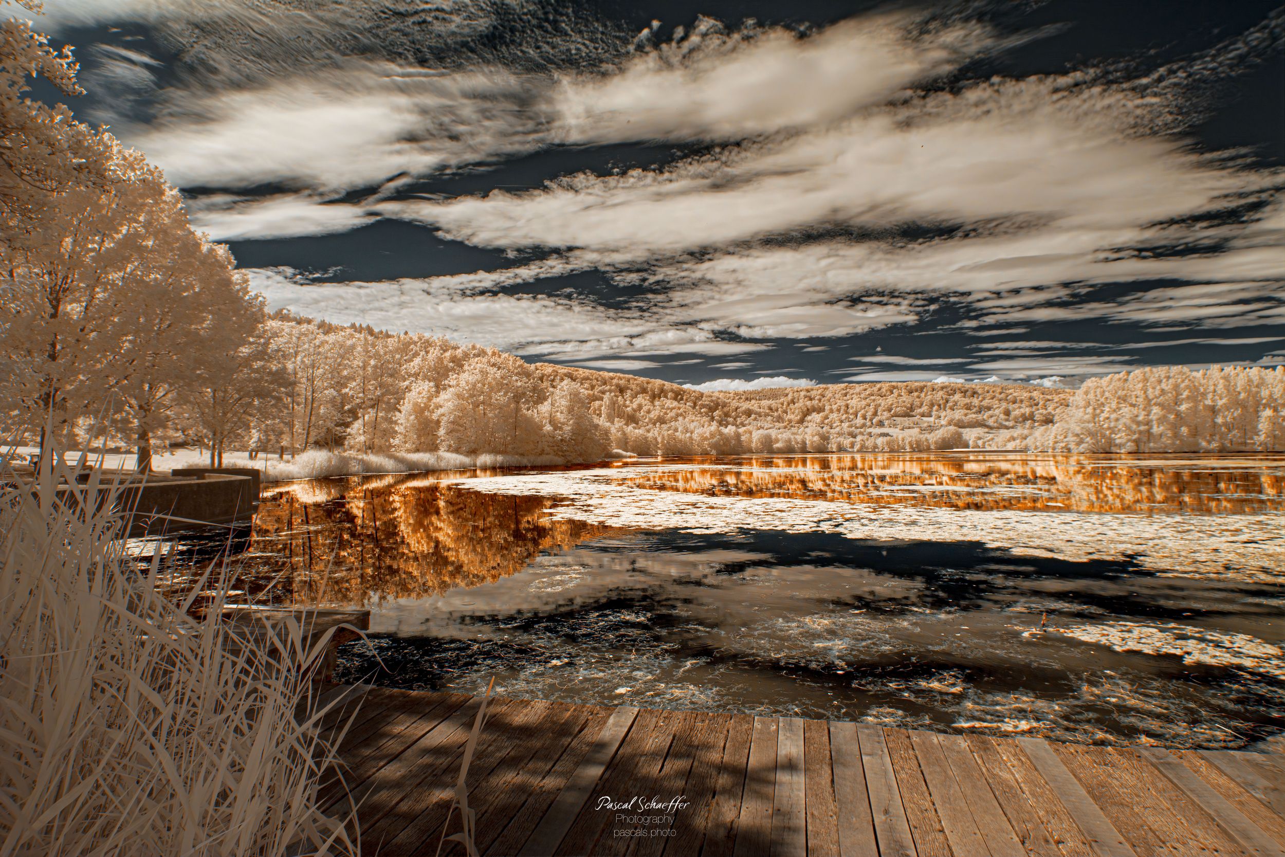 Infrared landscape photo of a serene lake reflecting golden trees under dramatic cloudy skies with a wooden deck foreground.