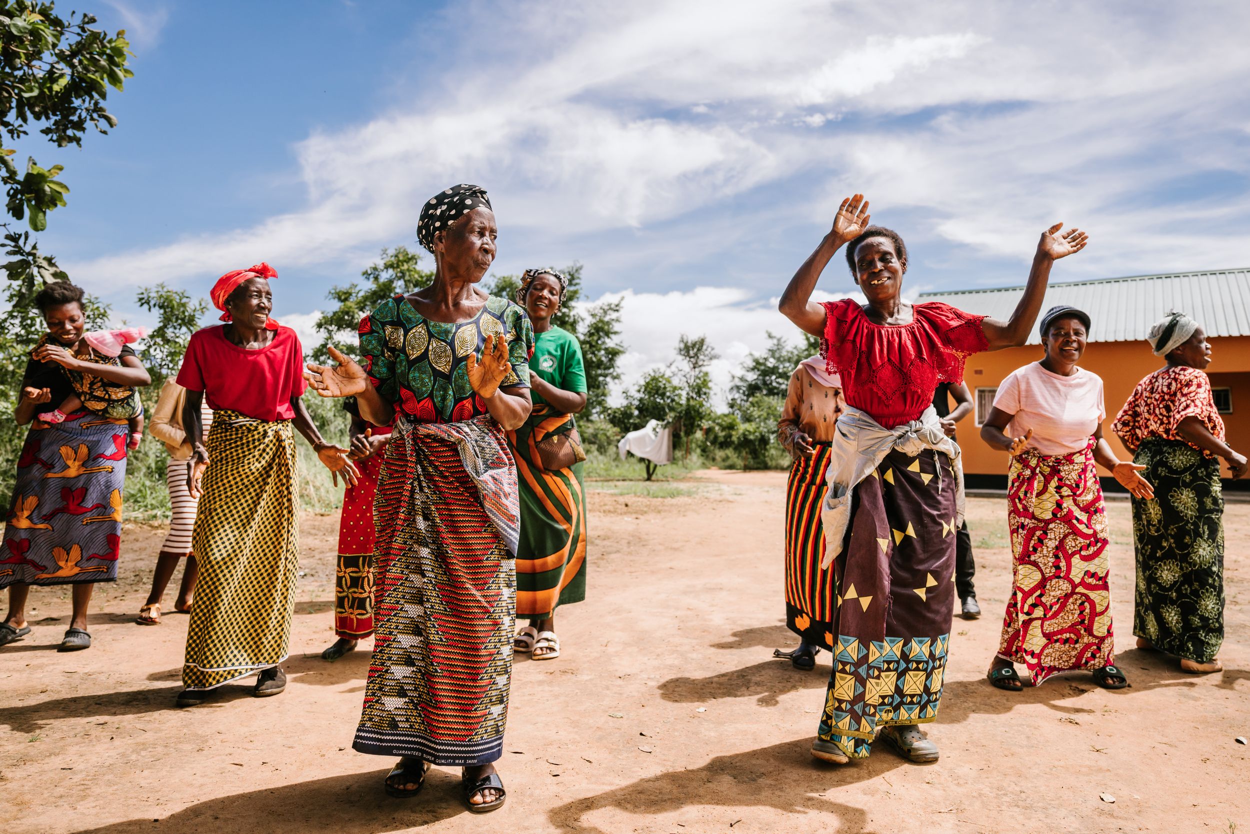 African women in colorful traditional clothing dancing outdoors on a sunny day in a rural village setting.
